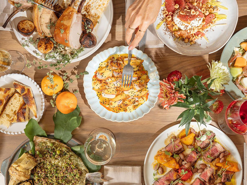Overhead view of a Mediterranean feast at Bastia, with shared plates including charred flatbread, grilled vegetables, citrus-dressed salads, sliced steak with stone fruit, fresh pasta, and artfully plated seafood.